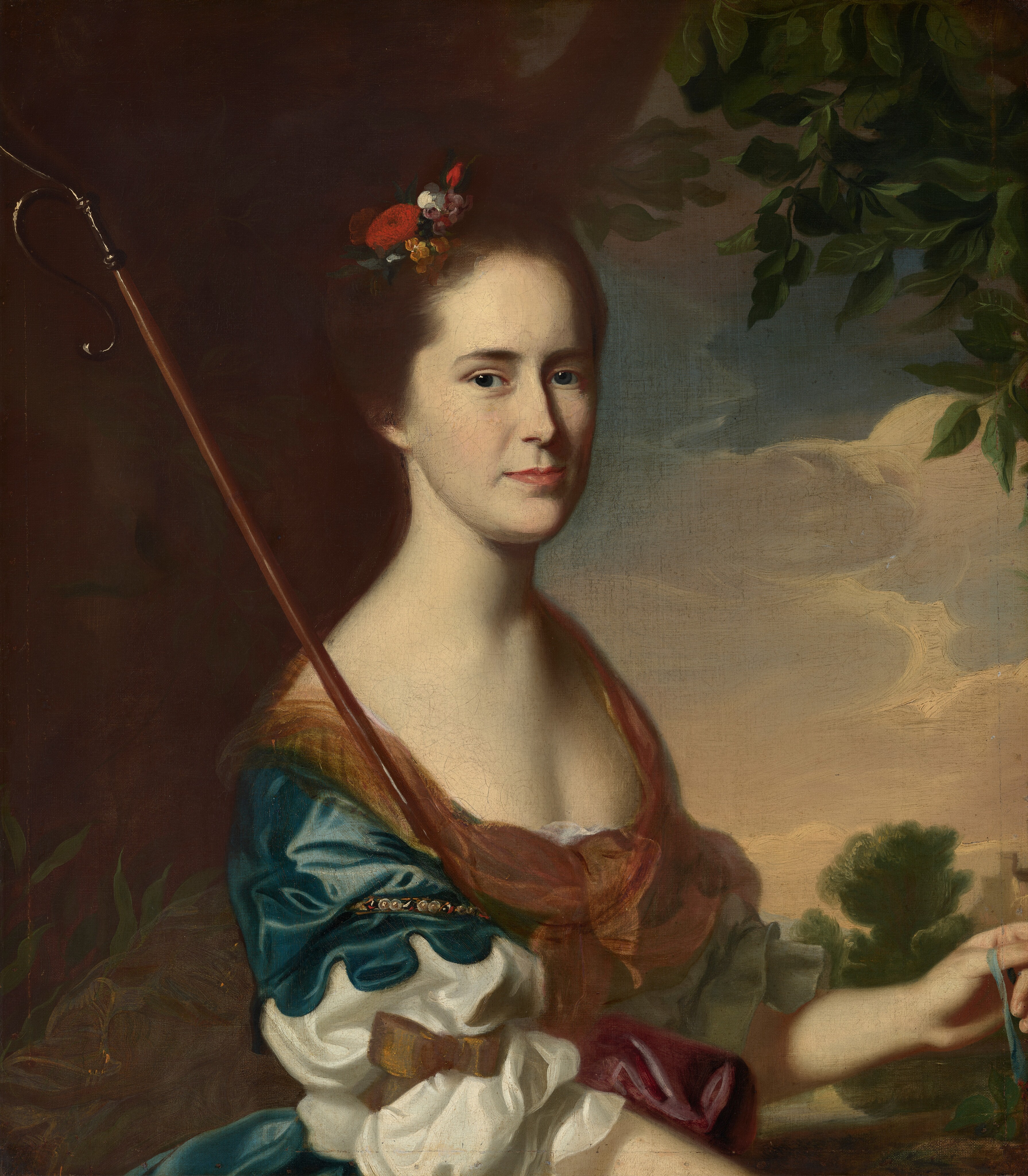 This painting is a portrait of a woman. She has fair skin, a straight nose, and thin red lips, with her dark hair styled up with flowers. She is wearing a multi-colored dress with voluminous sleeves, a wide neckline, and a sheer golden scarf. The woman is sitting and holding a hooked staff in one hand and a thin ribbon in the other. Behind her is a landscape background with a large tree on the left and a cloudy sky on the right.